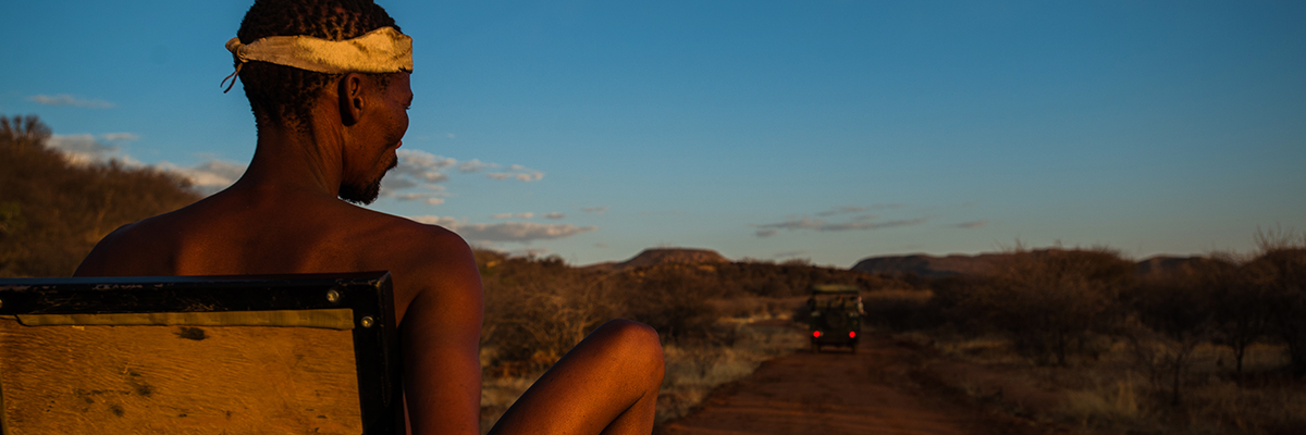 A San Bushman sits on a 4x4 seat overlooking the plains and another truck
