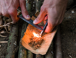 A knife scrapes across a ferrocium rod, causing sparks to fly into a bundle of tinder.